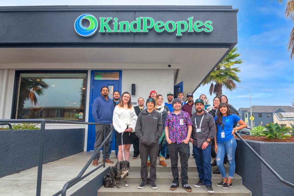 A group of smiling cannabis dispensary staff standing on the steps of the cannabis dispensary on a bright sunny day